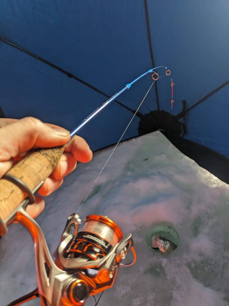 KastKing Kestrel ice fishing reel and rod in action with rod bent and line in ice hole with crappie fish caught and just coming out of ice hole